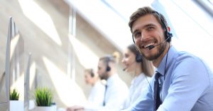 Customer support representative man wearing a headset and looking at the camera while the other two employees in the background working at their desk